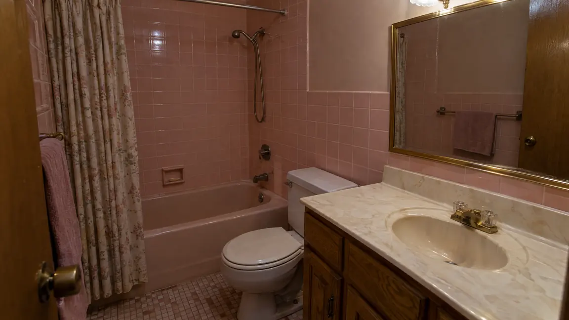 Before: Pink Tile Bath | Music City Remodels Before: A dated 1980s bathroom with pink wall tile, a cramped tub and shower combination, an oak vanity with a cultured marble top and a brass-framed mirror, dim lighting