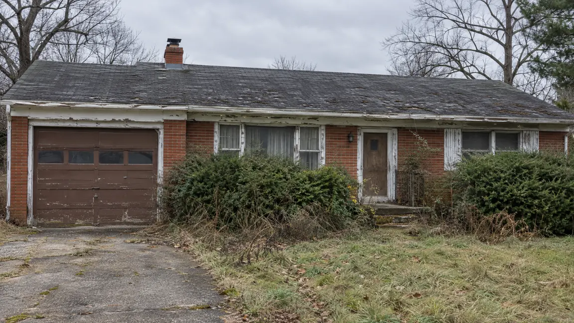 Before: Faded Exterior | Music City Remodels Before: A faded dated brick ranch house exterior with peeling white trim, an old brown garage door, a worn shingle roof, overgrown shrubs and an old front door under a gray sky